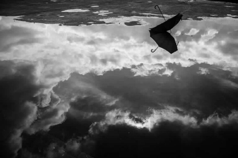 Scatto in bianco e nero di una pozzanghera su cui si riflette un cielo pieno di nuvole di diverse tonalità di grigio, ed in mezzo alla quale c’è un ombrello aperto, abbandonato, col manico rivolto verso l’alto (foto di Alicja Brodowicz).