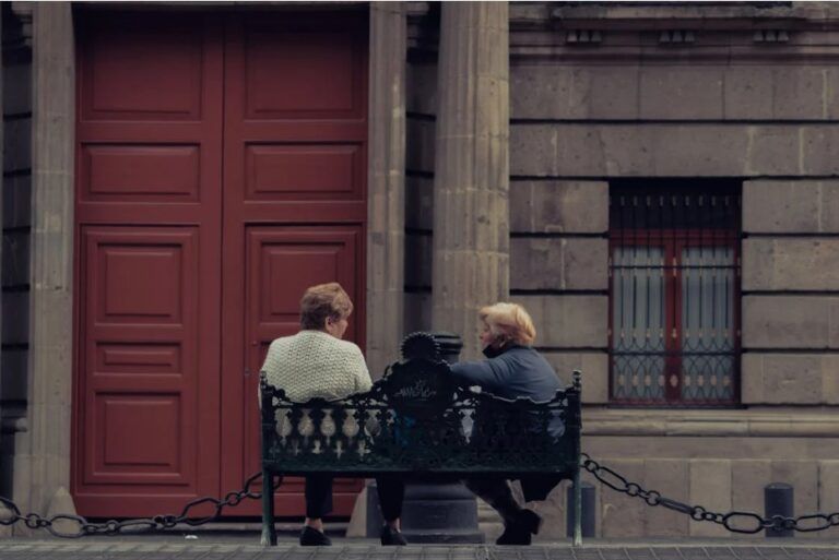 Due donne anziane, ritratte di spalle, sedute su una panchina, davanti ad un antico palazzo (foto di Josue Fuentes su Pexels).