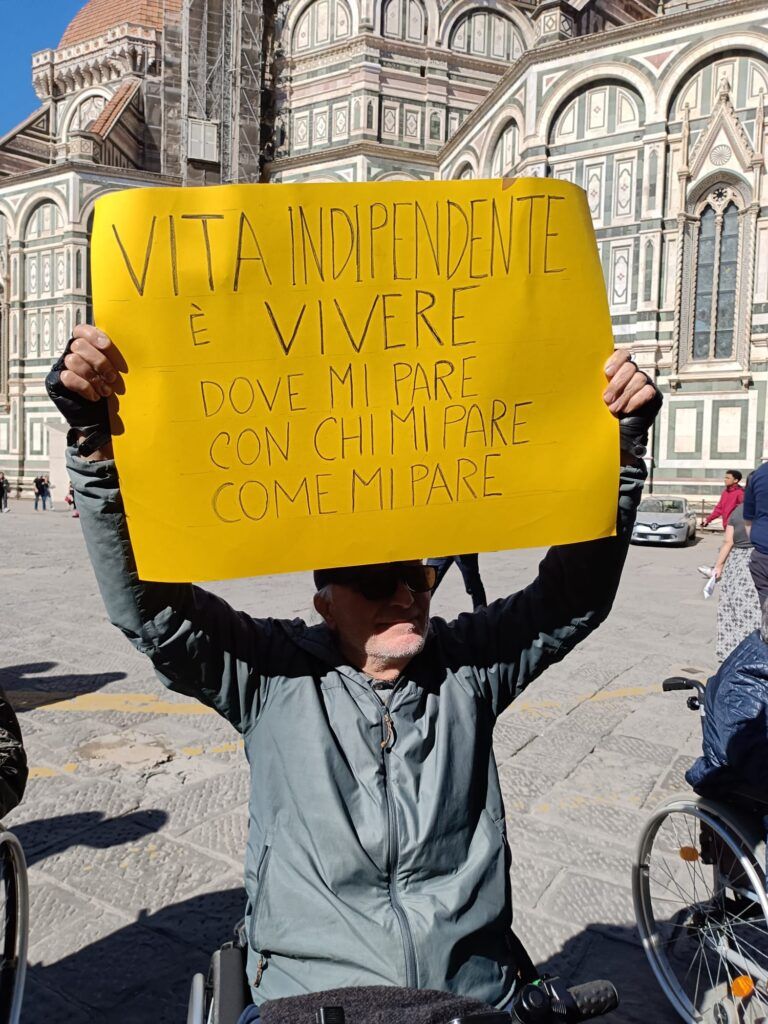 Un signore con disabilità, intervenuto al presidio di protesta indetto dall’Associazione AVI Toscana in difesa dei servizi per la Vita Indipendente, esibisce un cartello giallo con la scritta “Vita Indipendente è vivere dove mi pare, con chi mi pare, come mi pare (Firenze, 20 marzo 2024).