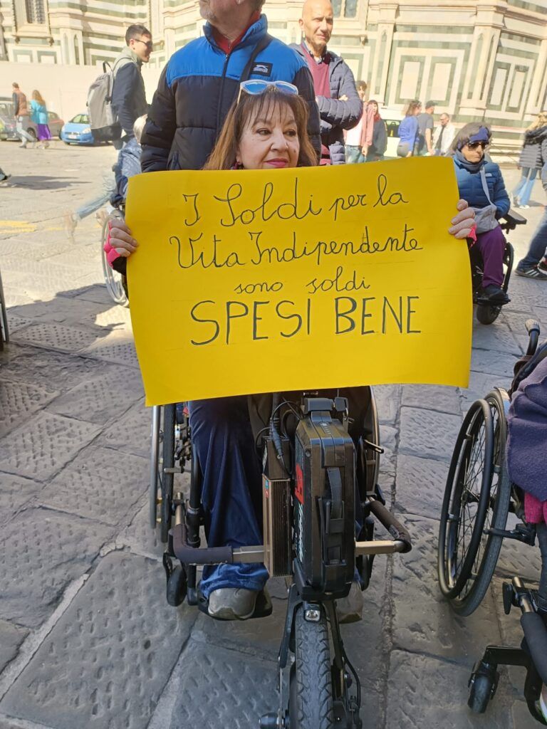 Una donna con disabilità, intervenuta al presidio di protesta indetto dall’Associazione AVI Toscana in difesa dei servizi per la Vita Indipendente, esibisce un cartello giallo con la scritta “I soldi per la Vita Indipendente sono soldi spesi bene” (Firenze, 20 marzo 2024).