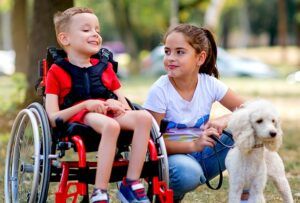Un bambino in sedia a rotelle e una bambina, in un parco, portano a spasso un cane bianco di piccola taglia.