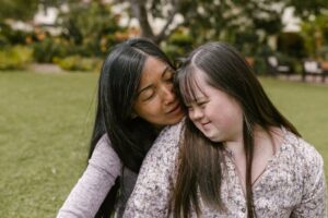 Una caregiver parla con una donna con sindrome di Down ponendosi dietro di lei e poggiando il viso sulla sua spalla in un gesto affettuoso (fonte: Pexels).