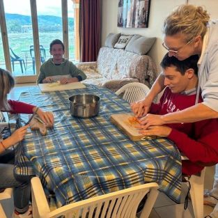 Attività di persone con disabilità alla “Casa di Mario” dell’Associazione Oltre lo Sguardo. Sedute attorno ad un tavolo con una pentola al centro, tre persone puliscono e tagliano delle verdure su dei taglieri. Un giovane è aiutato in questa operazione da una donna più grande.