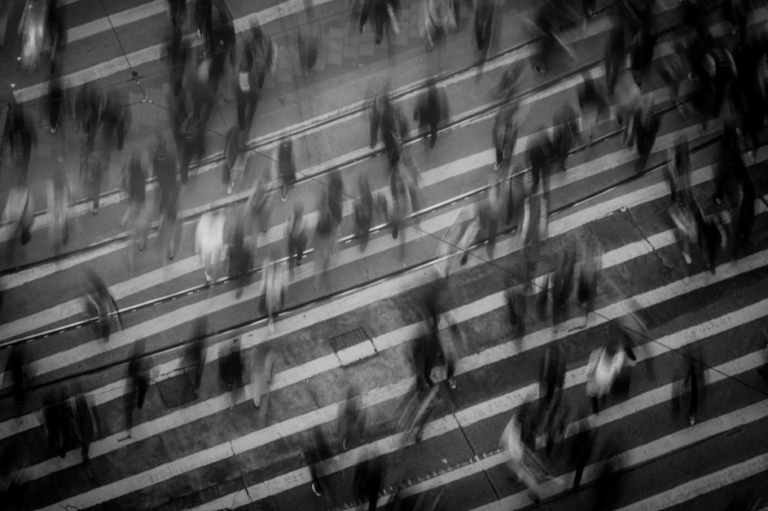 Sulle strisce di un enorme attraversamento pedonale si muove una moltitudine di persone sfuocate (foto di Mike Chai su Pexels).