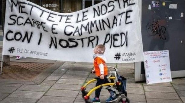 Un bambino con disabilità che si sposta con un deambulatore cammina per strada davanti ad uno striscione appeso da “Comitato Caregiver Familiari B1 B2 affondate”, sul quale è scritto: “Mettete le nostre scarpe e camminate con i nostri piedi”.