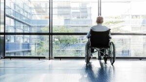 Un uomo in sedia a rotelle ritratto di spalle mentre guarda fuori da un’ampia vetrata.