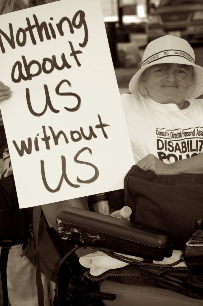 Foto in bianco nero di una donna con disabilità che manifesta per i propri diritti esibendo un cartello con la scritta “Nothing about us without us” (Nulla su di noi senza di noi).