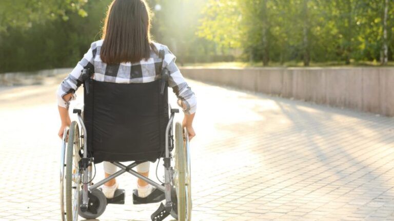 Una donna in sedia a rotelle ritratta di spalle mentre attraversa una piazza circondata da alberi in una giornata luminosa.