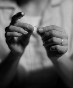 Particolare di due mani che infilano un filo nell’ago. In un dito è presente un ditale (foto in bianco e nero di Lee Friedman).
