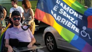 Simone Giangiacomi, orgoglioso, porta con sé una bandiera arcobaleno sulla quale è riportato il logo stilizzato di una persona in sedia a rotelle e la scritta “L’amore è senza barriere”.