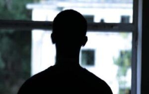 La parte superiore di un uomo, ritratto di spalle e in controluce, all’interno di una struttura, mentre guarda un edificio fuori dalla finestra.