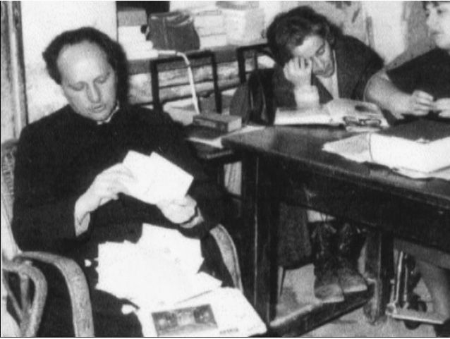 Un’immagine in bianco e nero ritrae don Lorenzo Milani mentre, seduto, consulta alcune lettere che tiene poggiate in grembo.