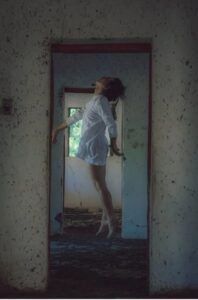 Una donna scalza e con un miniabito bianco spicca un salto all’interno di un ambiente fatiscente e senza infissi (foto di mododeolhar su Pexels).