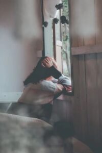 Una donna giovane ritratta seduta e di lato dentro una stanza con una parete di legno. La donna guarda fuori da una finestra e poggia i gomiti sul davanzale mentre tiene le mani nei capelli lunghi e castani (foto di Dương Nhân su Pexels).