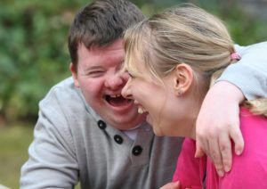 Un uomo con disabilità cinge col braccio le spalle della sua caregiver mentre ridono assieme.