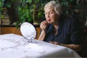 Una donna anziana si dà il rossetto guardandosi in uno specchio poggiato su un tavolo (foto di Anna Shvets, su Pexels).