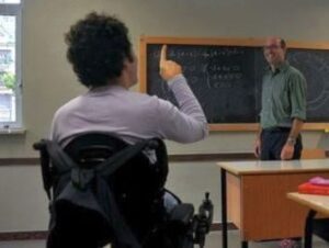 In una classe delle scuole superiori, uno studente in sedia a rotelle ritratto di spalle alza la mano per rivolgere una domanda all’insegnante che spiega qualcosa alla lavagna.
