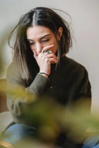 Una giovane donna seduta con le gambe incrociate poggia una mano sulla parte inferiore del viso con fare pensieroso (foto di Liza Summer su Pexels).