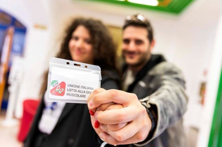 Una volontaria e un volontario della UILDM nostrano con orgoglio un tesserino identificativo dell’Unione Italiana Lotta alla Distrofia Muscolare.
